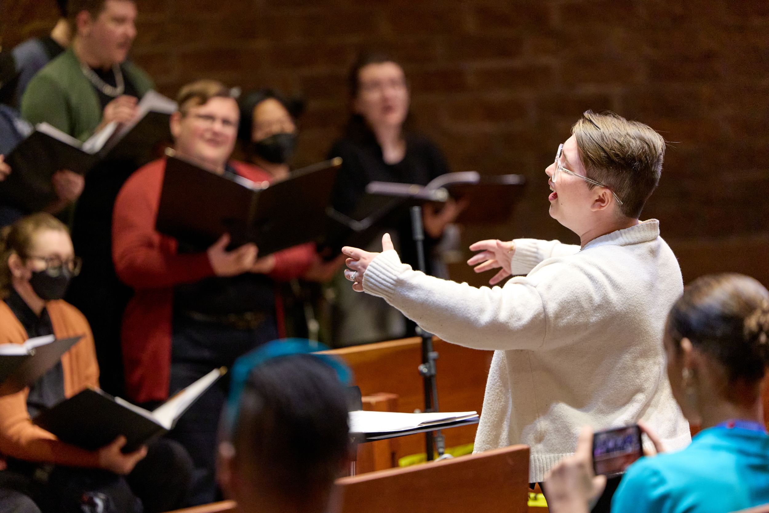 Closeup on a joyful choir conductor in a white sweater, black-clad choir members in soft focus beyond