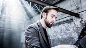 Closeup on Trifonov, a white man with brown hair, wearing a gray blazer, playing piano in a dramatic industrial building