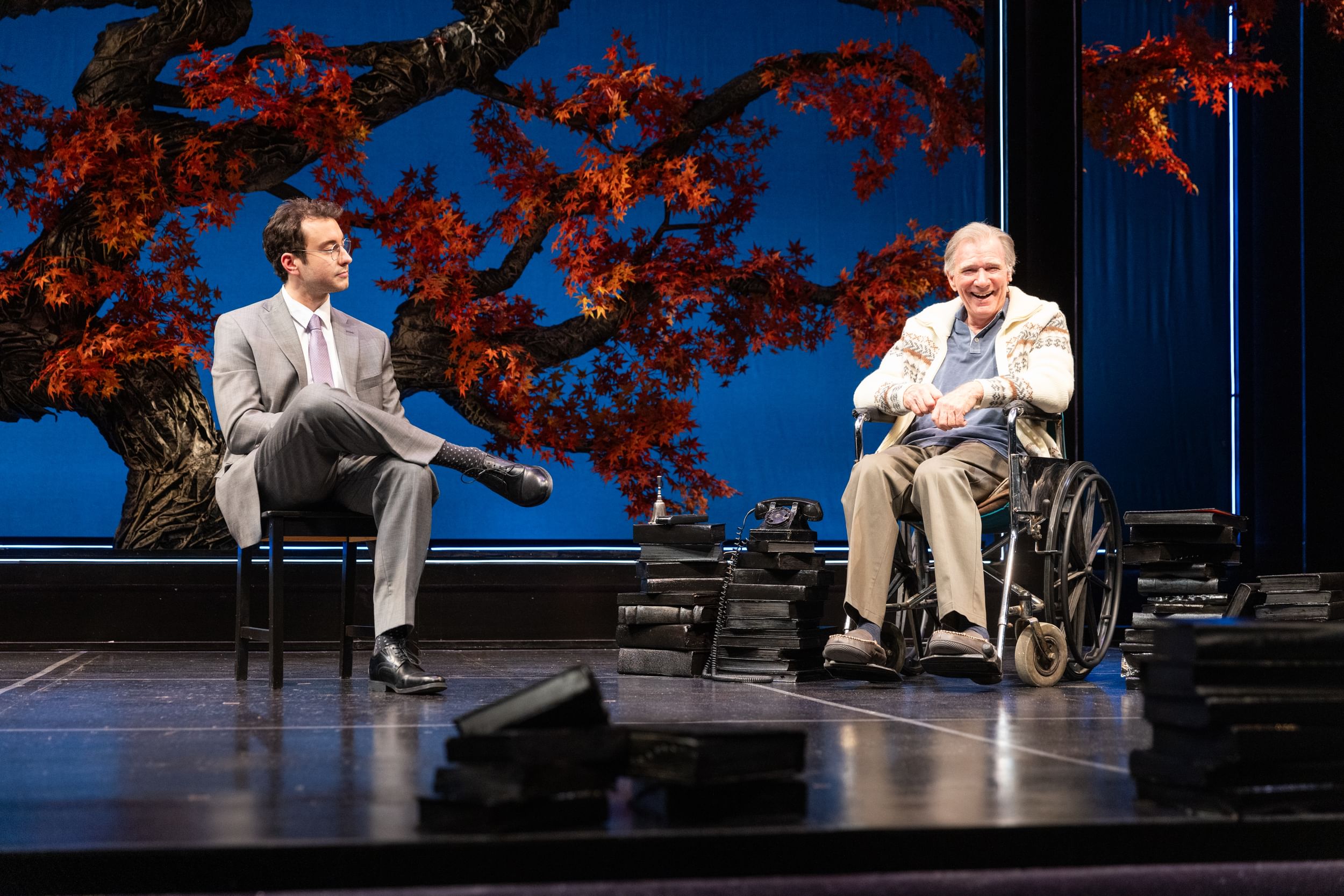 In front of a brilliant red Japanese maple, Howard sits in a chair and DeLaurier, smiling, sits in a wheelchair.