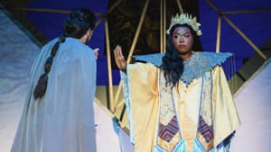 In opulent blue & gold robe, collar and stole, a frowning Johnson holds out her hand in a “stop” gesture toward Pickle.
