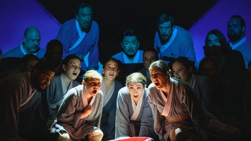 16 singers in gray robes crouch in a group onstage with a bright light shining in the middle of them.