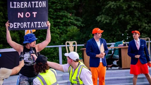 At left, an actor holds a sign reading LIBERATION NOT DEPORTATION in front of others in neon vests holding up mics.