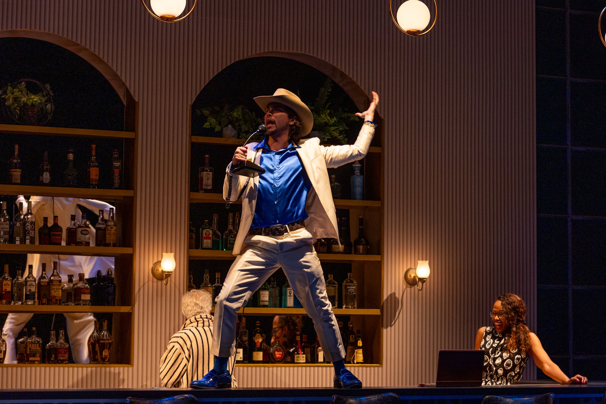 Lacey, in a cowboy hat, pale luxury suit, and blue shirt, sings wildly atop a hotel bar while Miles looks on grinning.