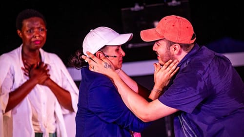Two actors in baseball hats embrace each other emotionally, while another looks on behind, her hands on her chest.