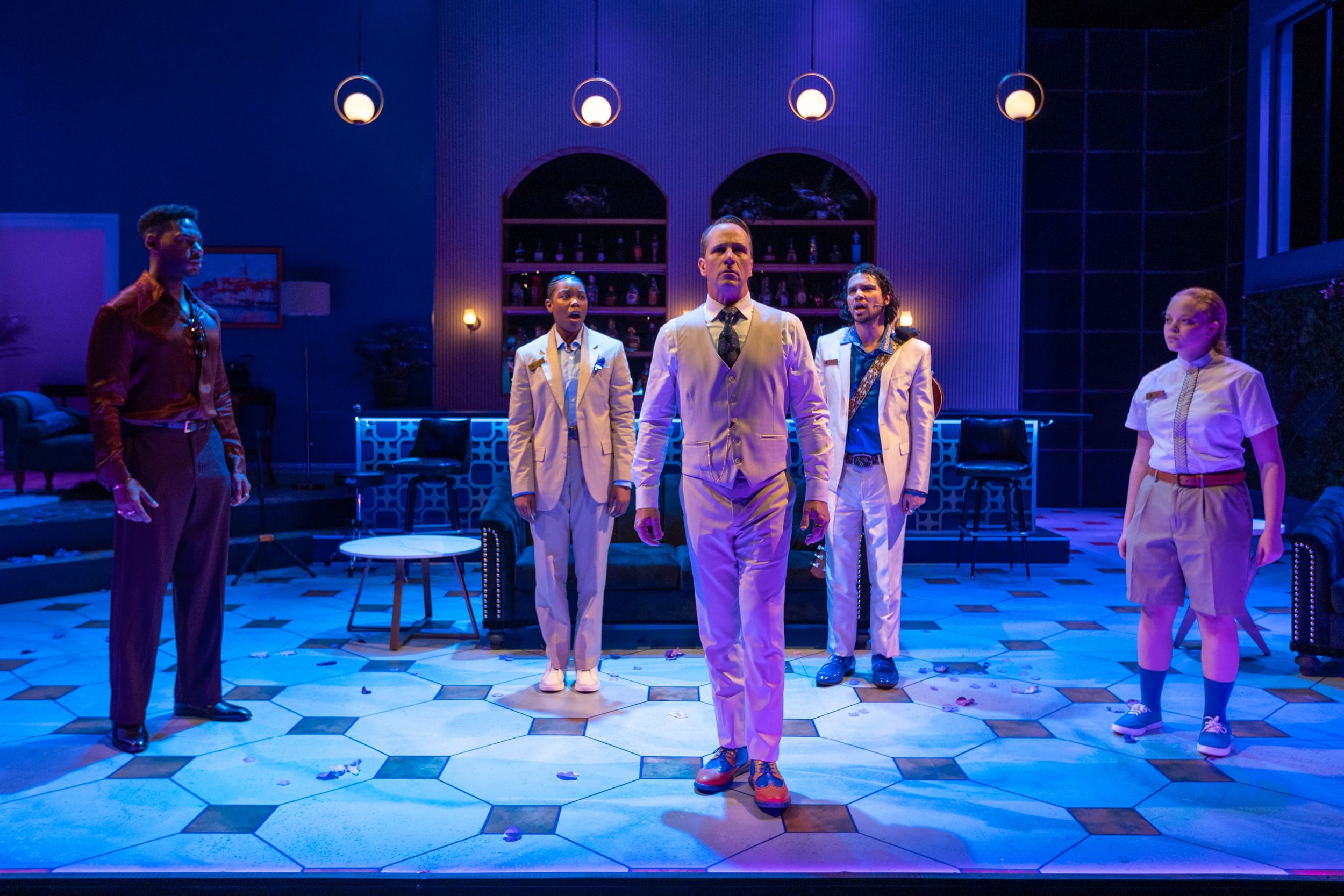 The ensemble, most of them in pale suits, pose together in dramatic blue light in a set like a ritzy hotel lobby.