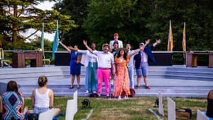 Ensemble of 8, in colorful costumes, poses together singing with arms outstretched, against a backdrop of green trees