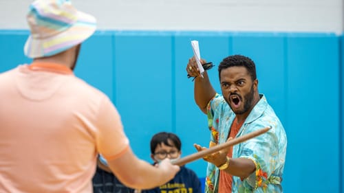 Buchanan, a Black man in tropical button-down, menaces Harrold with an exaggeratedly aggressive face, extending one arm.