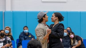 Photo of a performance in a community gym, with audience members wearing masks sitting around the two featured actors.