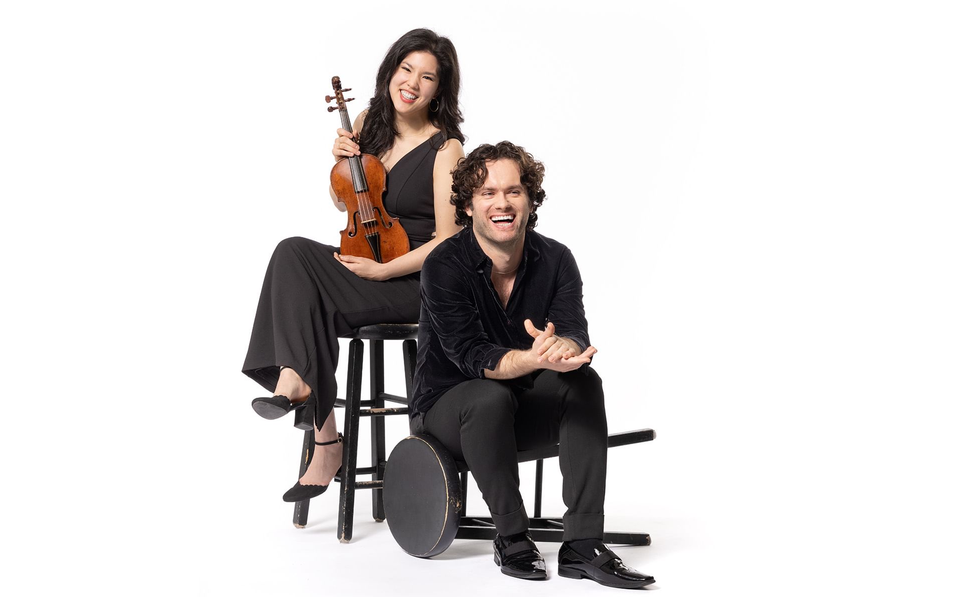 Portrait. Two performers sit on bar stool chairs in all-white room. Wong holds a violin.
