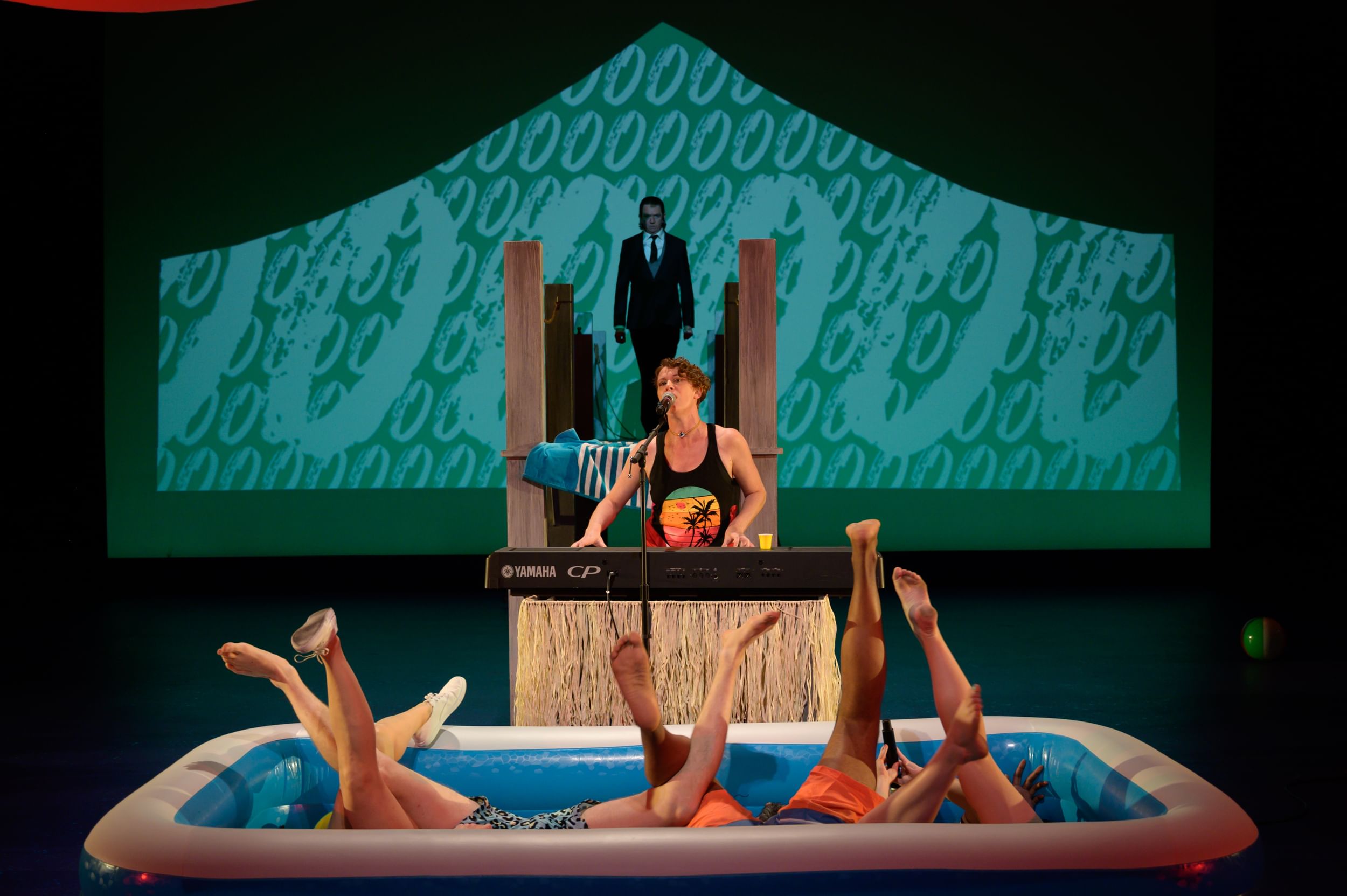 Conallen (Malvolio) paces on a pier above; Apple soulfully plays a keyboard; 4 sets of legs poke out intertwined from a pool