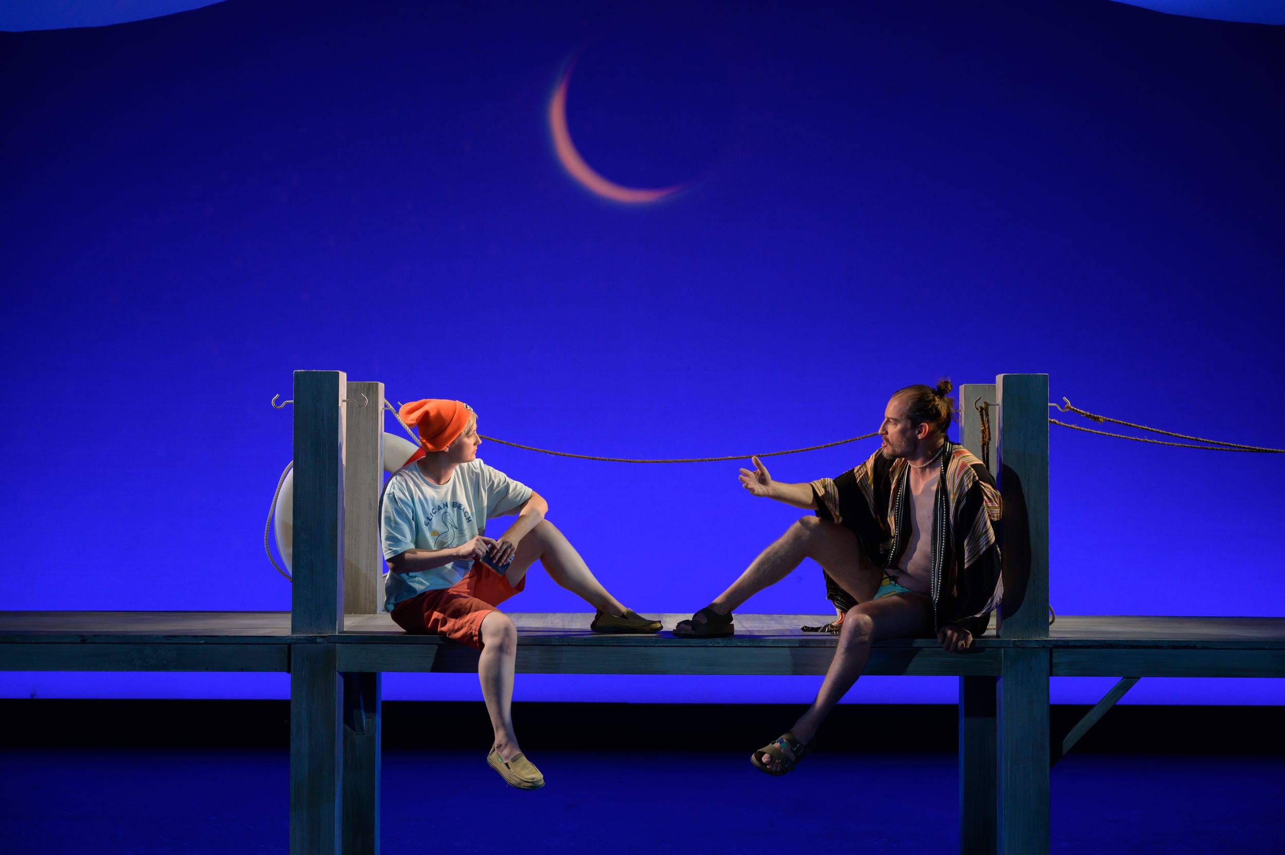 Tuomanen & Beschler mirror each other, leaning on the pillars of a pier in front of a rich, bright, night-blue backdrop