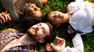 A Black woman, a white man, and an Asian person lie blissfully on the grass with their heads together and hands joined.