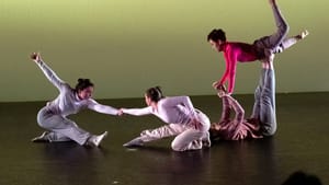 2 dancers on the floor reach to clasp the other’s hand. At right, one lies on her back, balancing another on her arms & legs