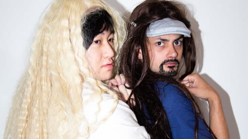 Shu wears a long blonde wig and Montalvo wears a long brown wig as they both look saucily over their shoulder.