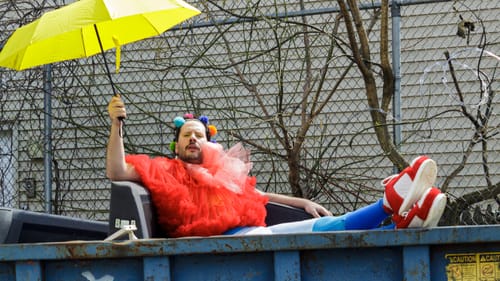 A man dressed like a clown, holding a yellow umbrella, reclines in a giant blue dumpster on a sunny day.