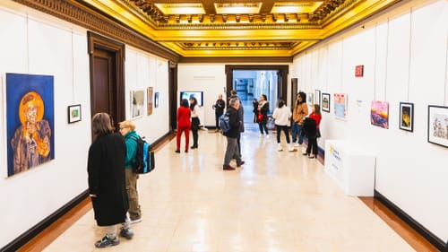 Bright photo of wide white City Hall corridor with gilded ceiling over the art on the walls and about 12 viewers.
