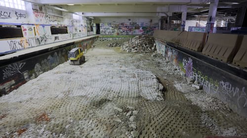A dry, empty pool with a torn-up cement floor, piles of rubble, and graffiti all around the surrounding abandoned room.