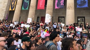 Hundreds of people, many holding handmade signs, rally on a June day on the steps of the pillared Hamilton Hall building.