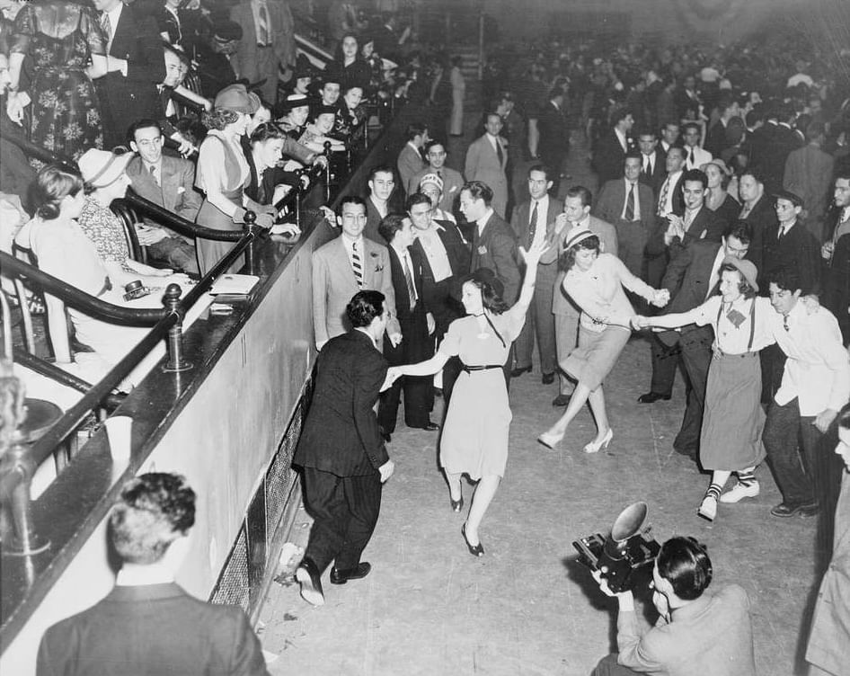 Vintage black & white photo of packed, joyful scene of 1940s revelers dancing, watching, and chatting in suits and dresses.