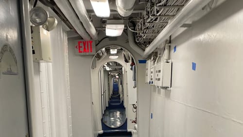 The blue floor, white wall, and series of doorways with rounded shapes in a hallway below deck recede straight away