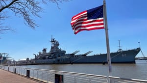 Exterior view of the huge ship on the river under a bright blue sky. An American flag flies in the foreground.