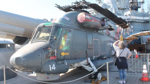 A woman, small next to a gray rescue helicopter on the ship deck, and other massive gear, looks up with her back to camera