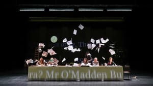 6 actors with exuberant expressions sit behind a long table onstage while many pages swirl through the air above them.