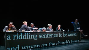 At a long table with text projected onto it, 7 actors of different ages, races, and genders sit in variously zany attitudes