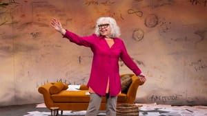 Grody, wearing a magenta blouse, in front of an orange chaise lounge and scattered papers, throws out her right arm & smiles