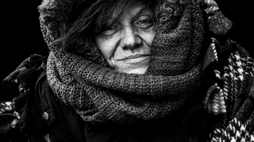 High-contrast photo of a slightly smiling woman with a weathered face, wrapped in bulky hoods and scarves.