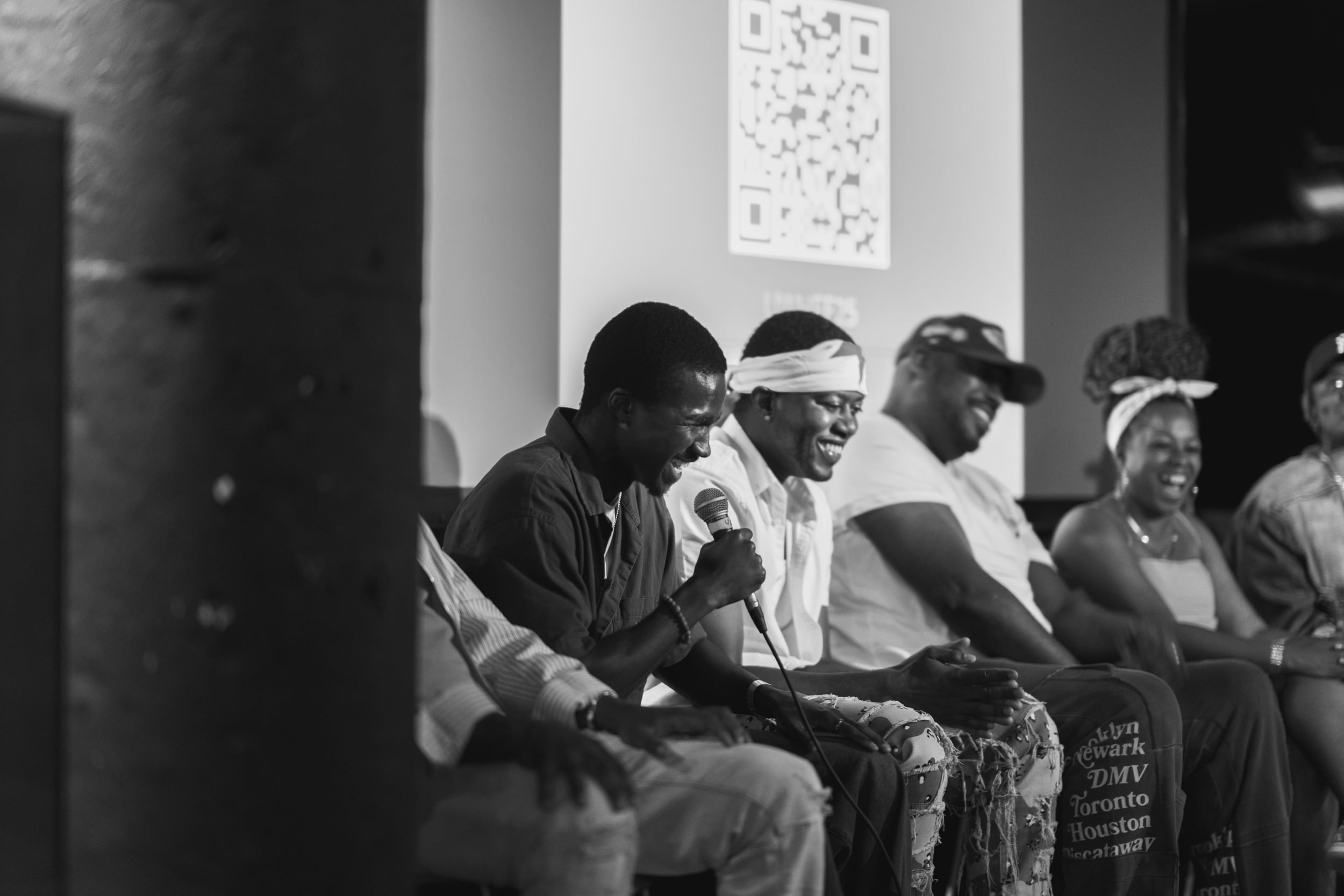 Black and white photo of about six people on stage for what looks to be a panel, in collective laughter. One holds the mic.