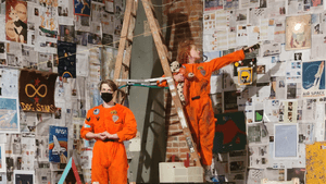 Sturtridge and Stoller, in orange flight jumpsuits, pose by a ladder in a room covered with media clippings and artwork.