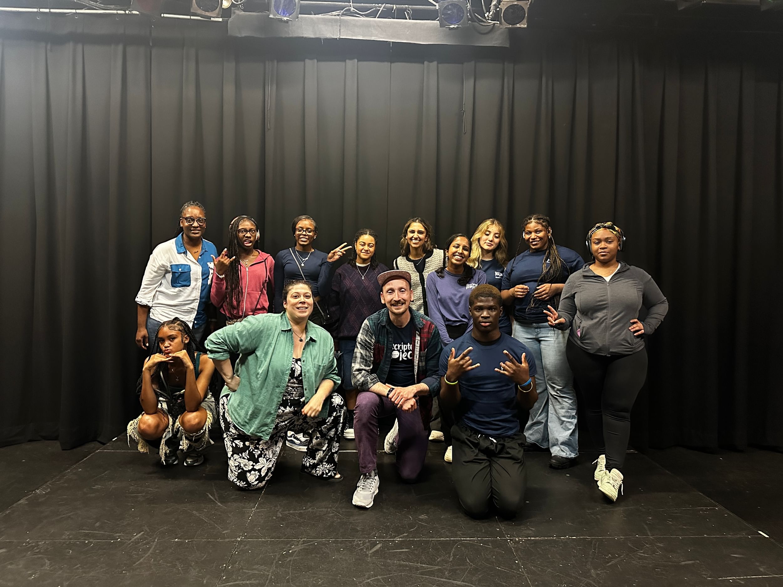 A group of teens and adults pose for a camera in a small black box theater space