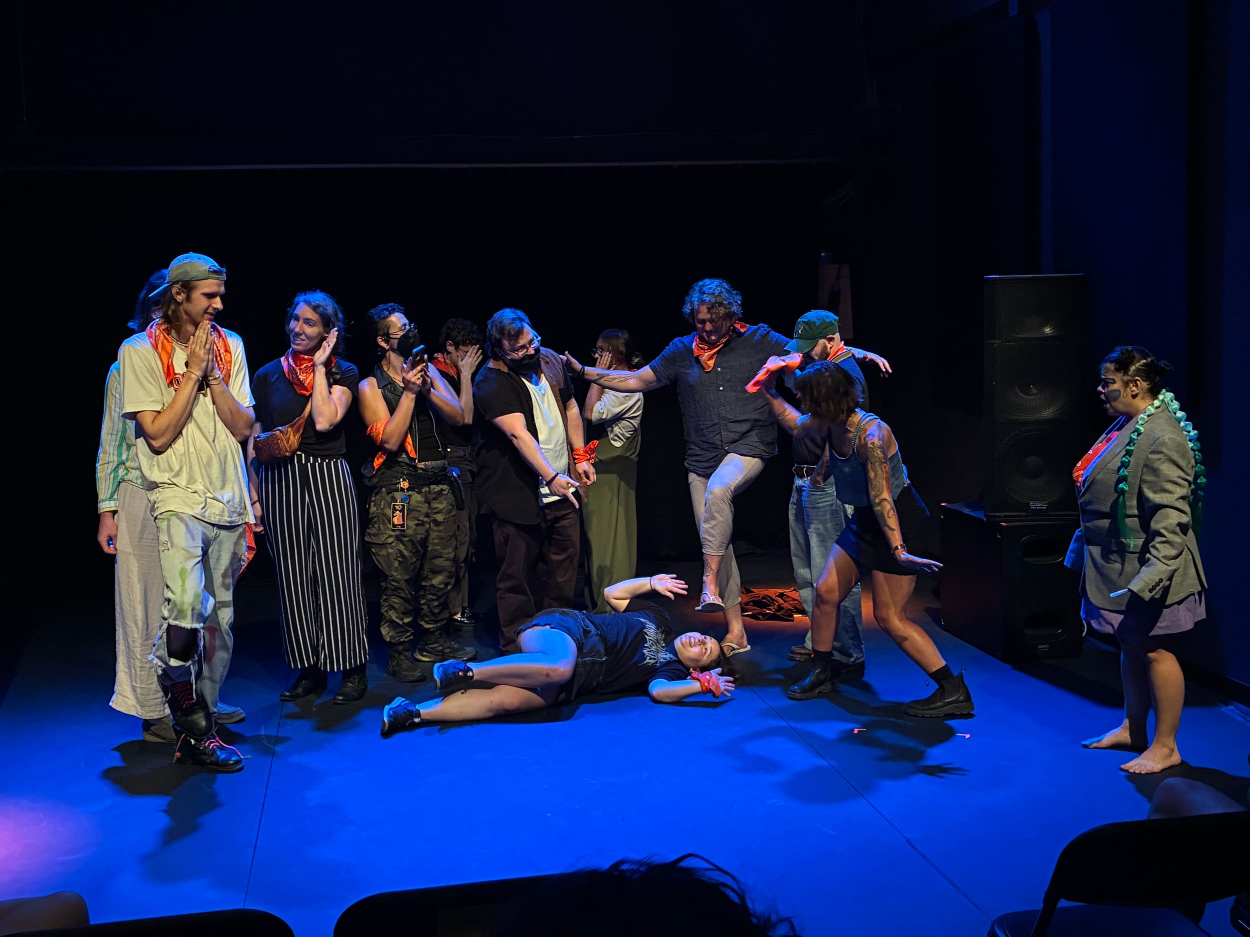 11 audience participants in casual clothes pretend to attack one on the ground at center, as Mender watches in horror