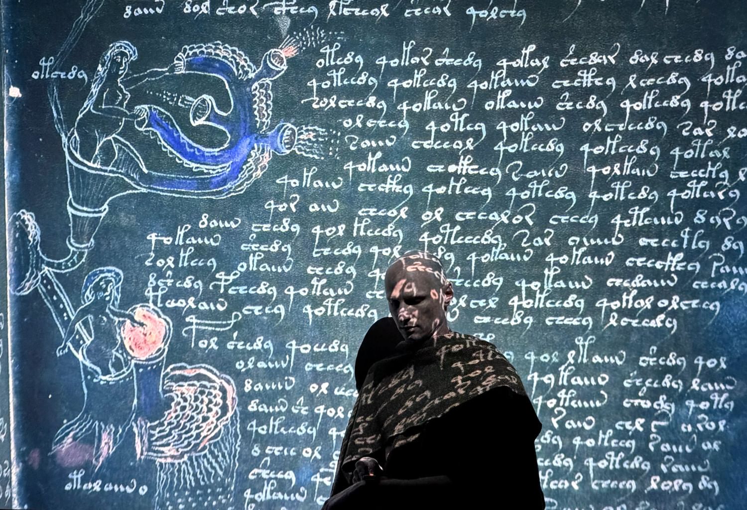 Gillette, a bald white man in a brown robe, poses in a projection of an illuminated Medieval manuscript in unknown language