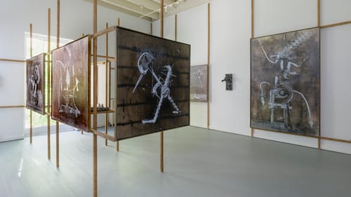 In an airy white gallery, large rectangular brown-and-white prints show vaguely anthropomorphic or animalistic machines.