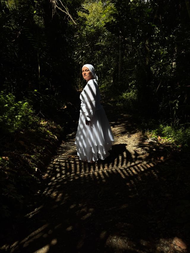 Woman in all-white stands at the center of tropical foliage. Shadows of palms cross her body.