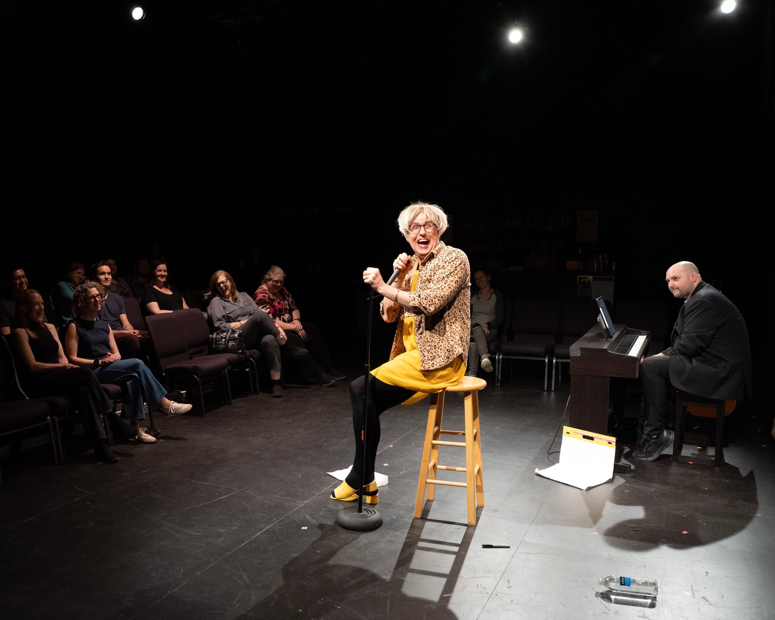 Kitrosser, in modest yellow drag with a leopard-print shirt, smiles joyfully at the mic in front of a small theater audience.