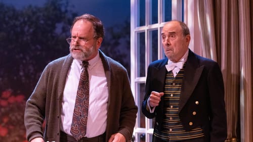 Scene from the show. Ingram, with a cardigan and gray beard, stands by the liveried Novelli, who looks disapproving.