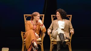 Scene from the play: sitting in adjacent forward-facing chairs, the two women turn their heads to look fondly at each other.