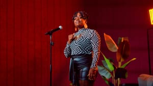 A Black woman in lace tights, black leather shorts, and a black-and-white checked blouse stands proudly at the mic.