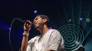 Nance, a Black man wearing a white shift, sits on a stool onstage and sings into a handheld mic, a spiral in light behind him