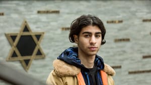 A young Iranian Jewish man faces the camera, straight-faced, the Star of David on a wall behind him.