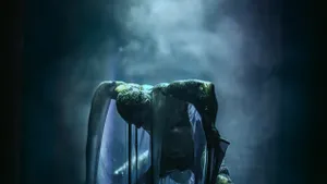 In a dramatic misty blue spotlight, a man rises animalistically, bent over with elbows angled up out of a well of viscous goo