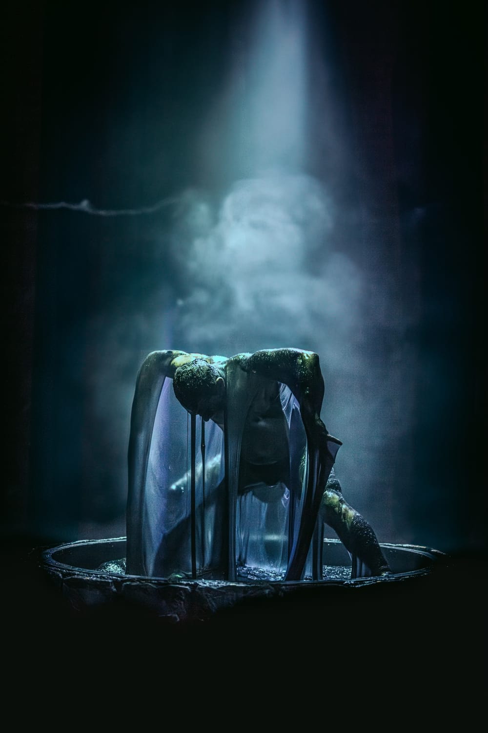 In a dramatic misty blue spotlight, a man rises animalistically, bent over with elbows angled up out of a well of viscous goo