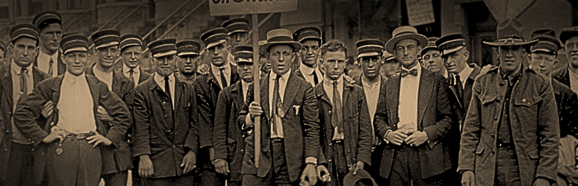 Sepia-toned vintage photo of men in suits, ties, hats. One in the center holds a sign, all in calm protest