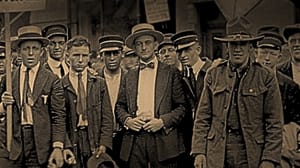 Sepia-toned vintage photo of men in suits, ties, hats. One in the center holds a sign, all in calm protest