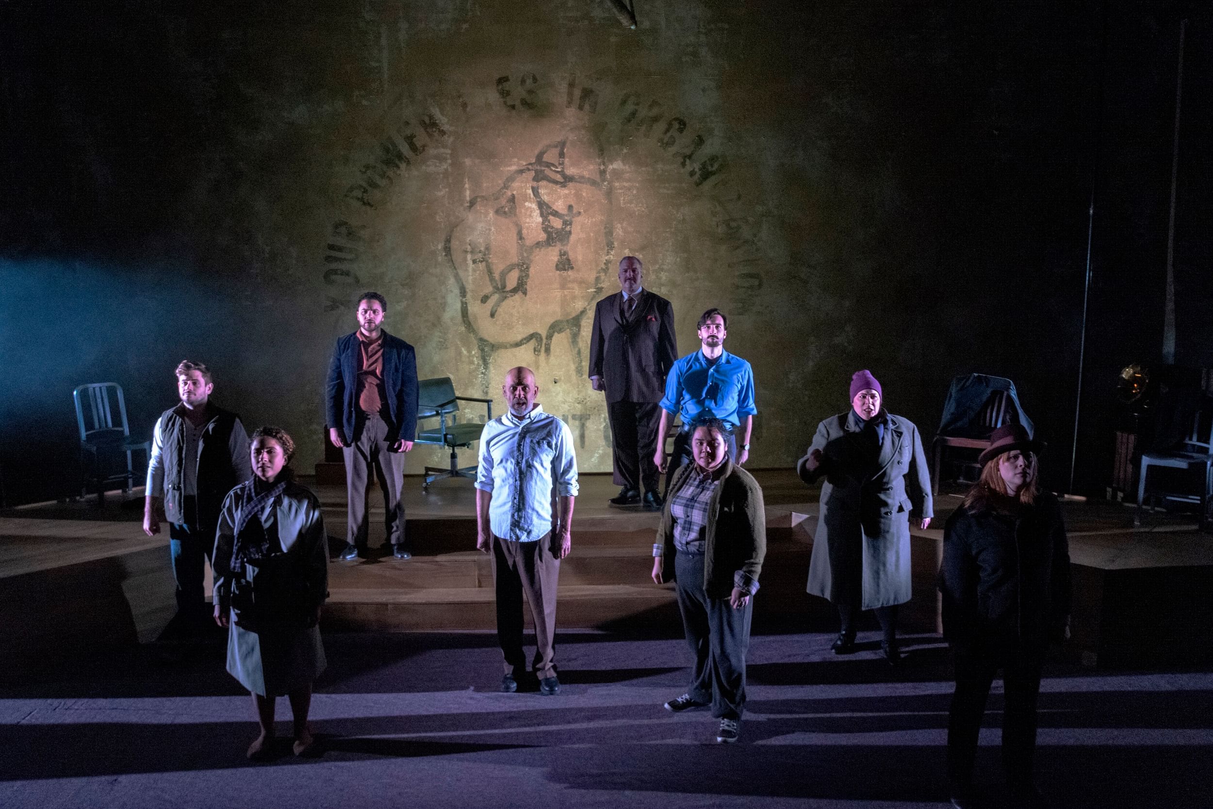 Nine cast members stand in a balanced arrangement facing the audience, on a wooden dais stage with low steps.