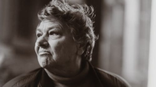 A black and white photo by Judith Joy Ross. It shows a solemn short-haired woman in her 50s. She wears a heart-shaped pendant