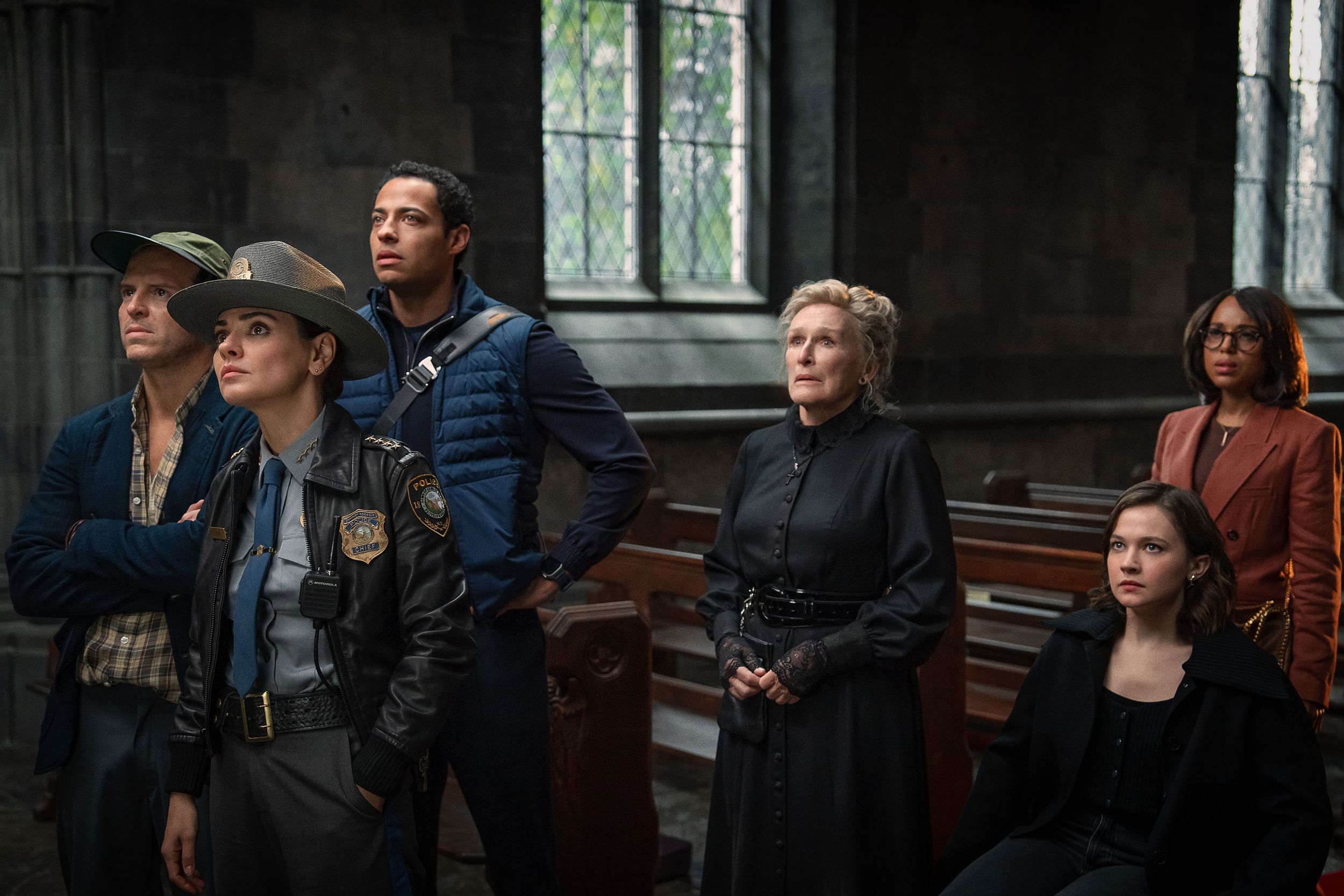 Six diverse actors stand with inquisitive, worried, or skeptical expressions in the wooden pews of an ornate church.